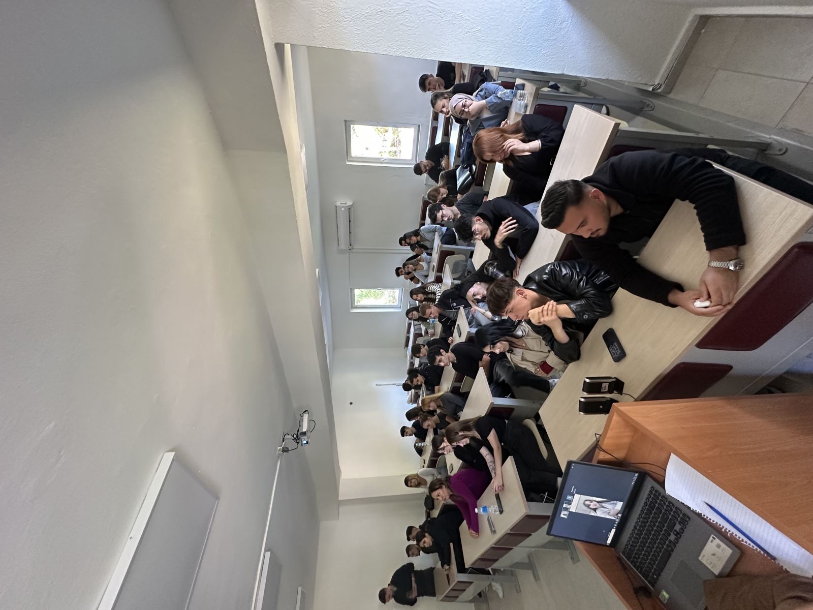 This photo shows the classroom from a wider angle. Around thirty university students are seated at desks, all facing forward and listening attentively. Some rest their hands under their chin, while others lean on their notebooks. At the front, a laptop is open on a podium, displaying a woman speaking in a virtual meeting. The classroom looks orderly, and the students appear highly focused on the speaker.